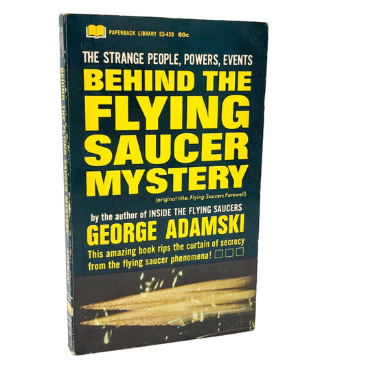The image displays the paperback edition of "Behind the Flying Saucer Mystery" by George Adamski, first published in 1967. The book's cover features a striking, vivid illustration of a flying saucer in mid-flight, with a backdrop of a stormy night sky.