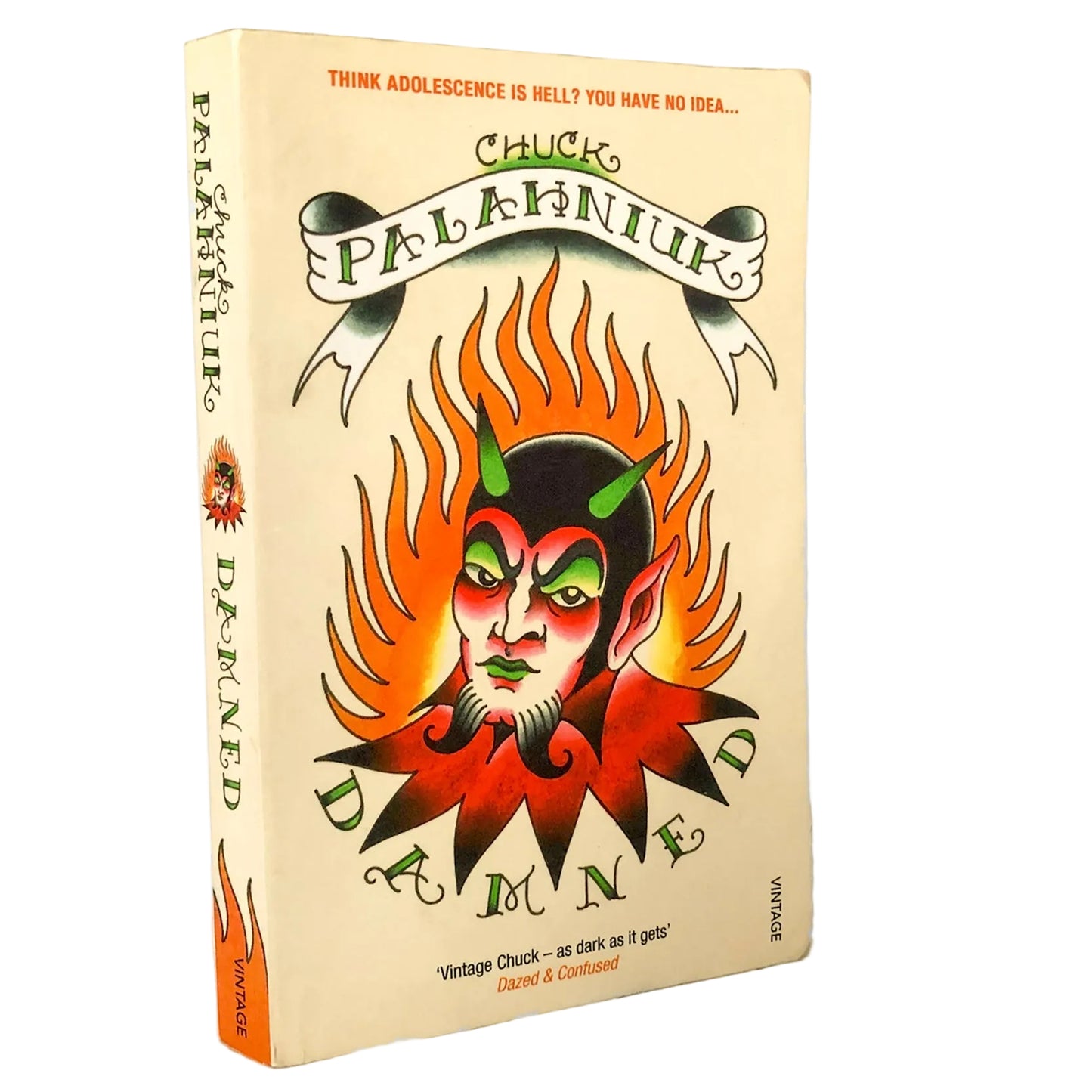 A paperback copy of the book 'Damned' by Chuck Palaniuk, standing upright on a marble-like surface against a white background. The cover features a cream-colored background with a central illustration of a demon face surrounded by orange flames, with the title 'DAMNED' in stylized green lettering.