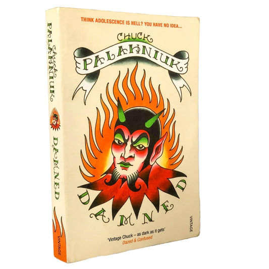 A paperback copy of the book 'Damned' by Chuck Palaniuk, standing upright on a marble-like surface against a white background. The cover features a cream-colored background with a central illustration of a demon face surrounded by orange flames, with the title 'DAMNED' in stylized green lettering.